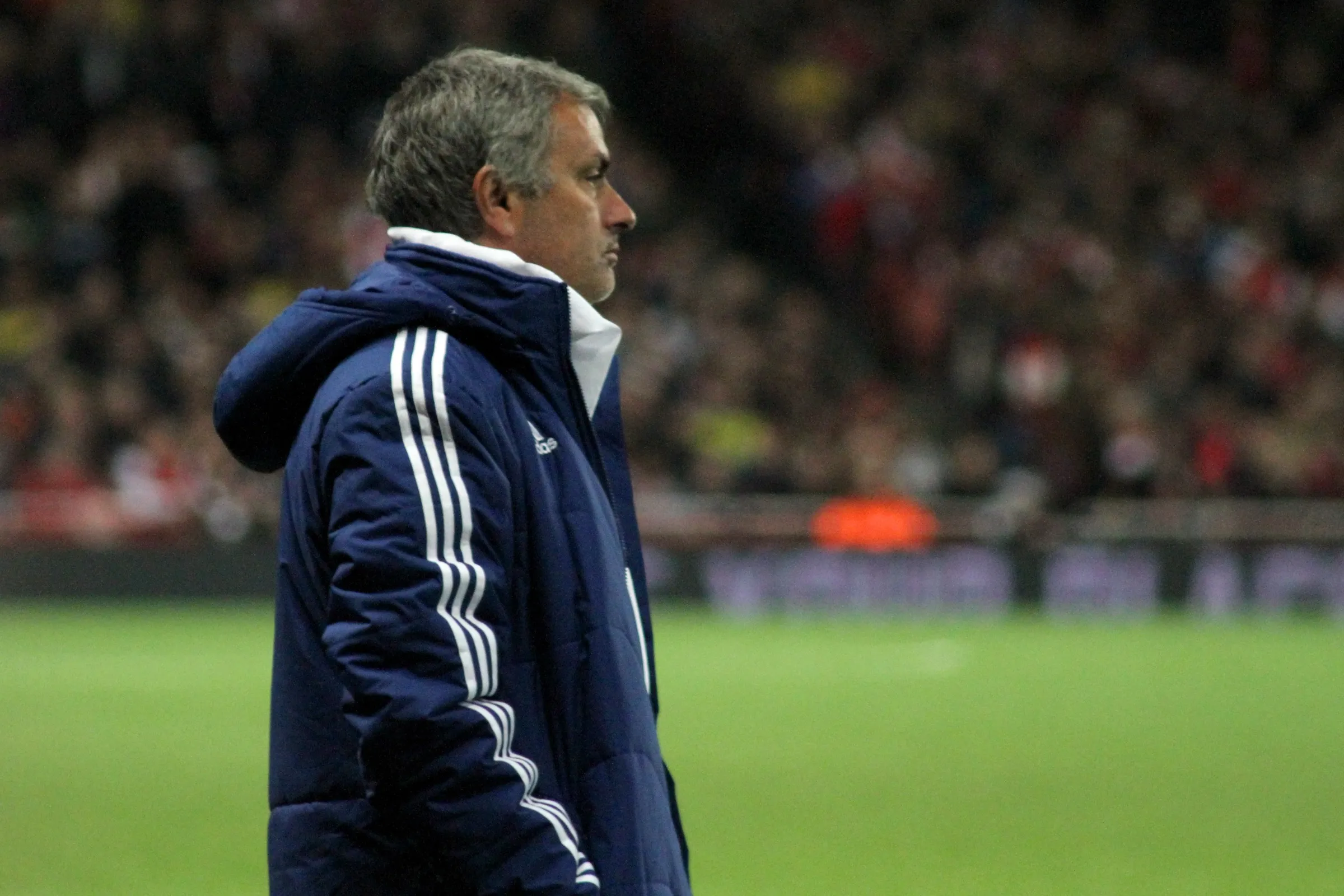 Jose Mourinho in a navy Chelsea coat, shot in profile on the touchline, staring out at a floodlit pitch.