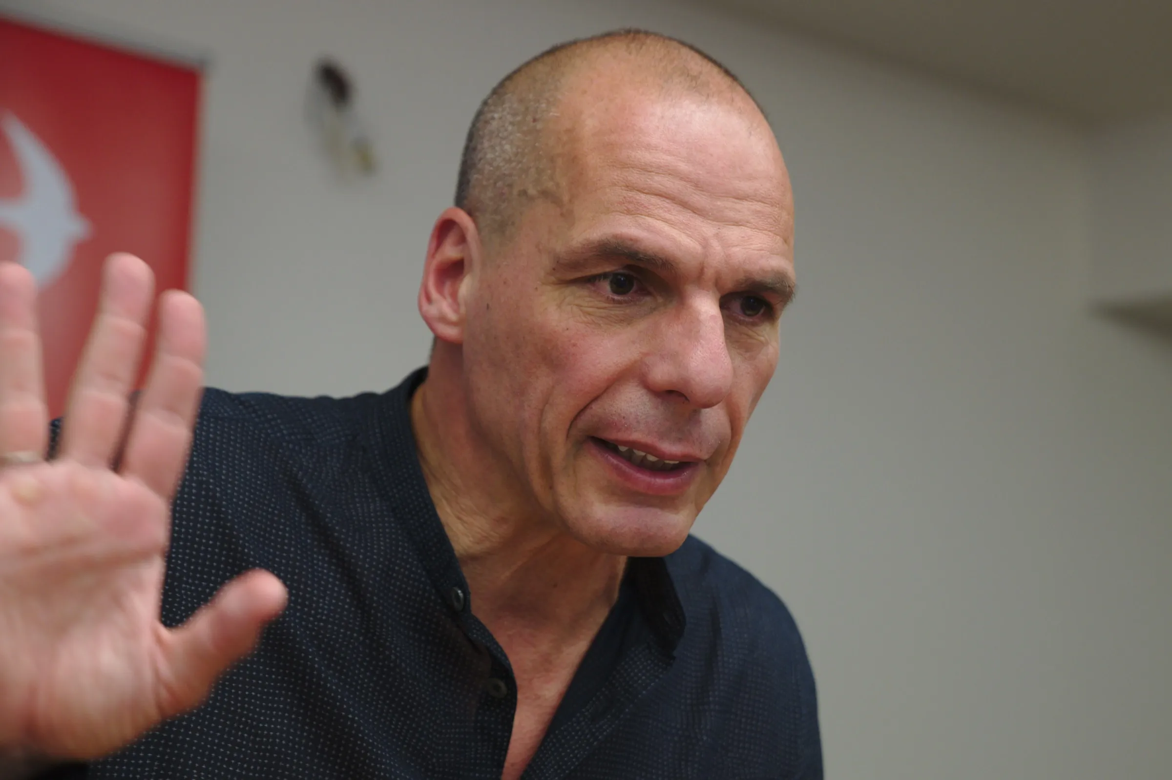 Yanis Varoufakis mid-sentence with a hand raised toward the camera, a red banner behind him.